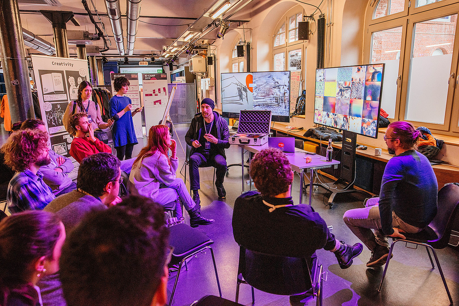 Eine Gruppe junger Menschen bei einem Workshop, sie unterhalten sich und schauen auf zwei Screens im Hintergrund