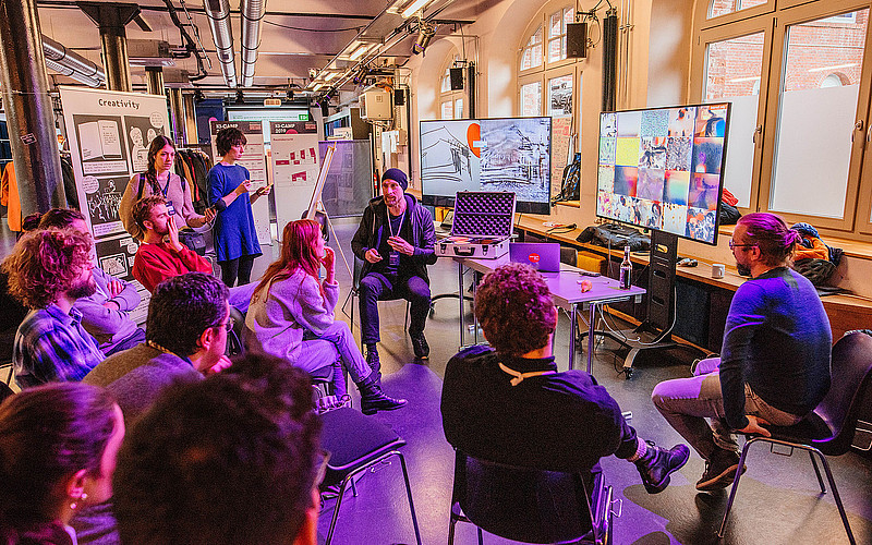 Eine Gruppe junger Menschen bei einem Workshop, im Hintergrund sind mehrere Screens zu sehen.